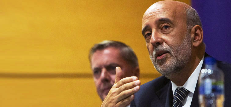 Governor Gabriel Makhlouf wearing a suit and tie pictured speaking with one hand raised with one person to his right. A yellow and blue wall is behind the two men.